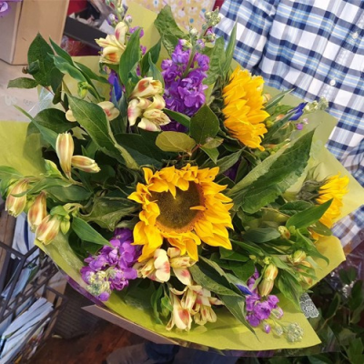 Hello Sunshine - A lovely hand tied bouquet tied and ready to go straight into a vase of water. This bouquet is created using the freshest blooms of the day in bright yellows and purples alongside complementary foliage. Created by an expert florist at Lily Blossom Florist in Huddersfield and delivered with care. A perfect bouquet to say Get well, Congratualtions, Happy Birthday or Thank you. Picture shows Sunflowers, scented Stocks, spring Iris and VERY long lasting Alstromeria (checked shirt not included)! A beautiful mix of dutch and english flowers. Why not purchase a vase alongside and we can deliver the flowers all ready to go, no need to do anything, just keep an eye on the water level. We always love anything with Sunflowers in - definitely a Hello Sunshine kind of vibe